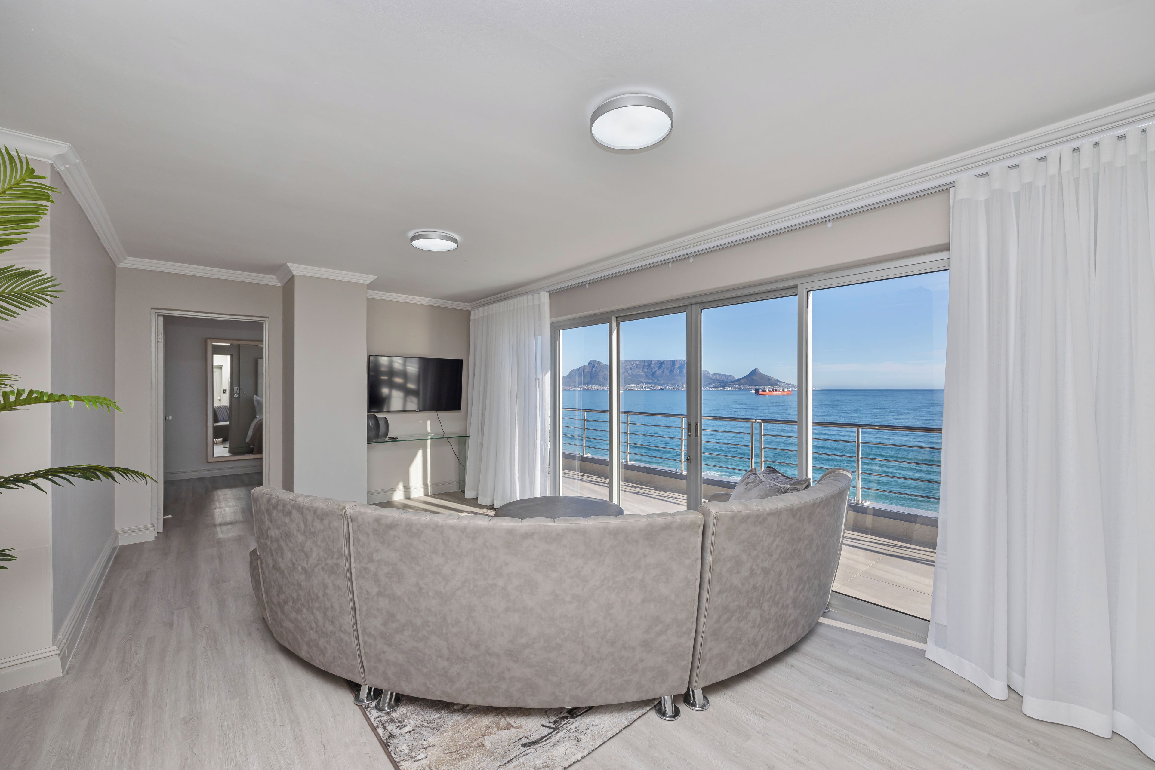 A modern living room with a curved grey sofa, a flat-screen TV, and a large window offering a view of the sea and mountains.