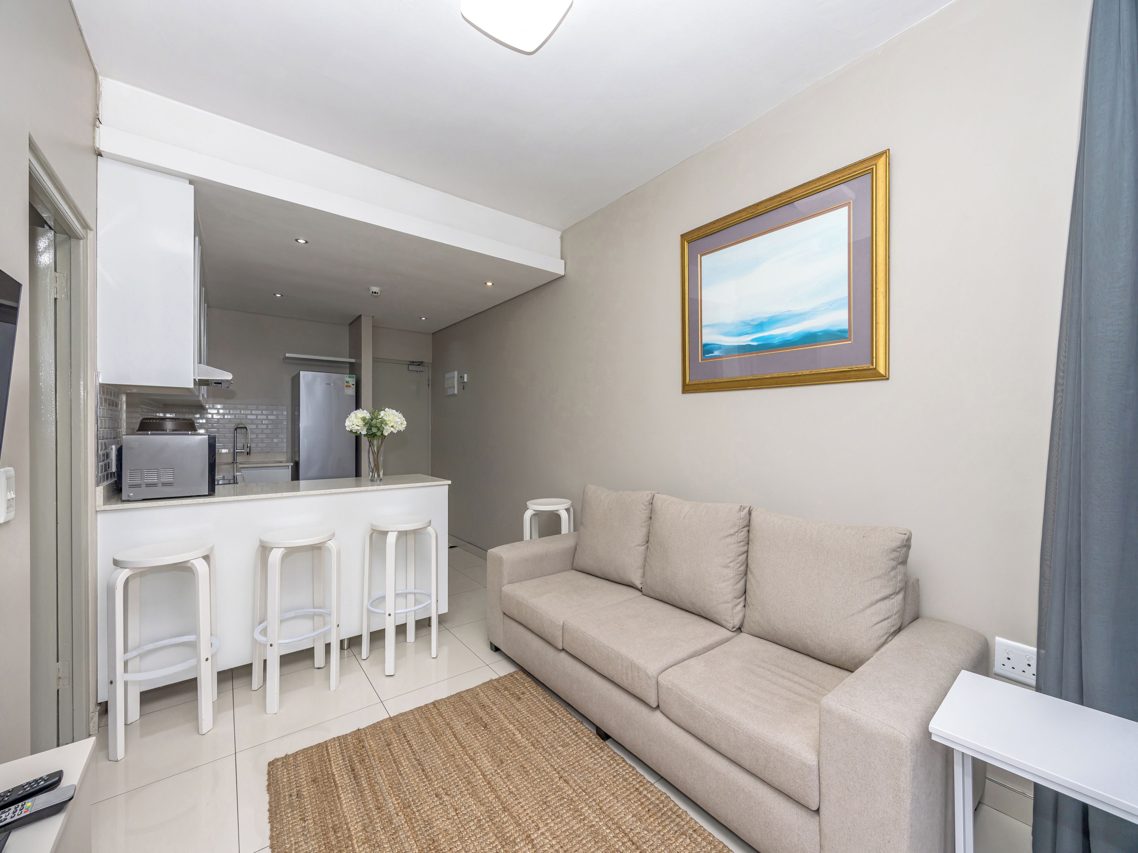 Una sala moderna con un sofá, una mesita blanca y una cocina con refrigerador y tostadora.