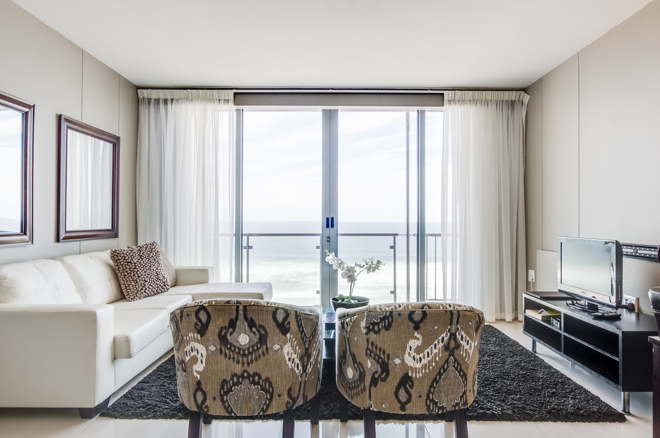 A modern living room with a sofa, two patterned chairs, a TV, and a large window with curtains.
