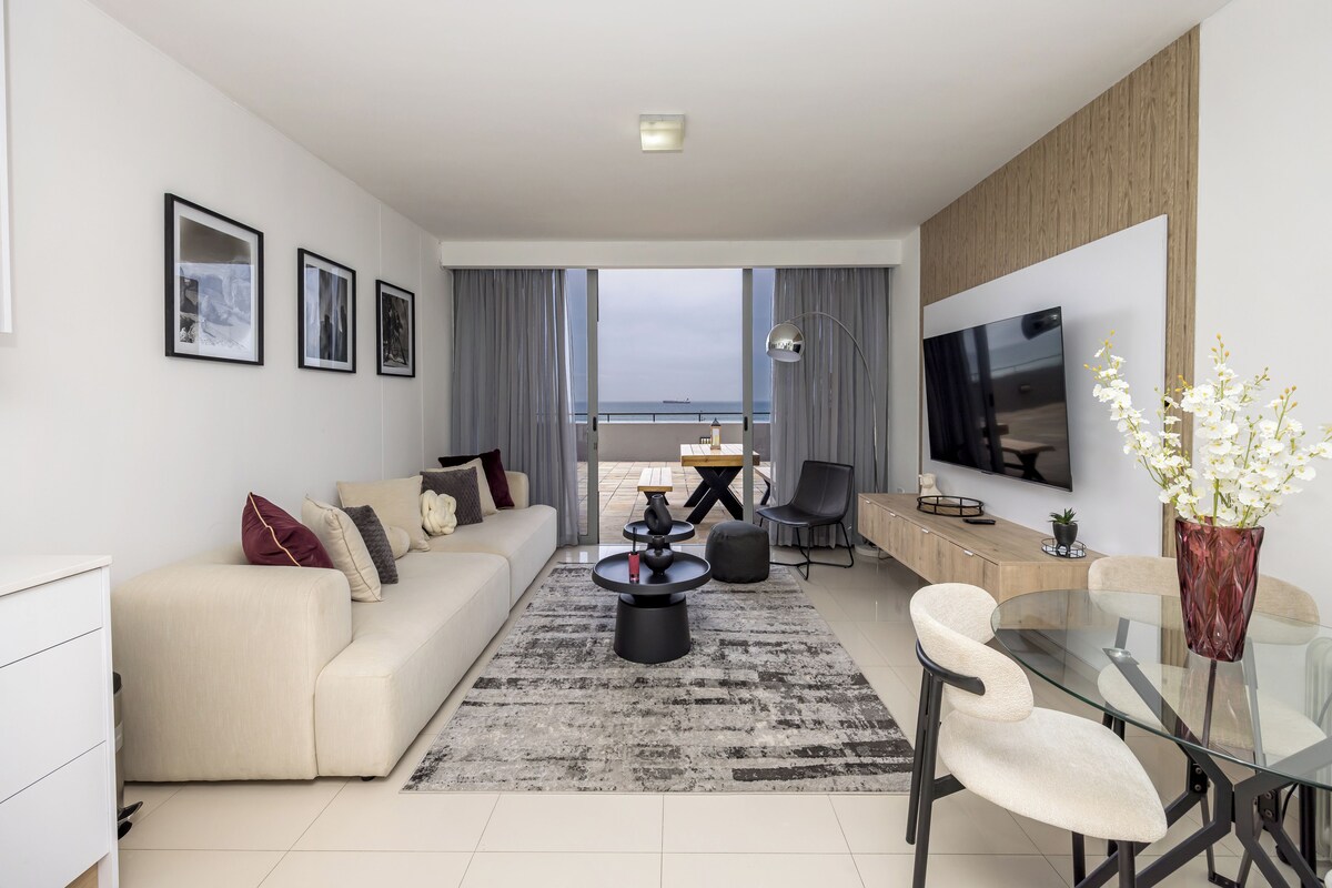 A modern living room with a sofa, coffee table, and TV. There is a balcony with a view.