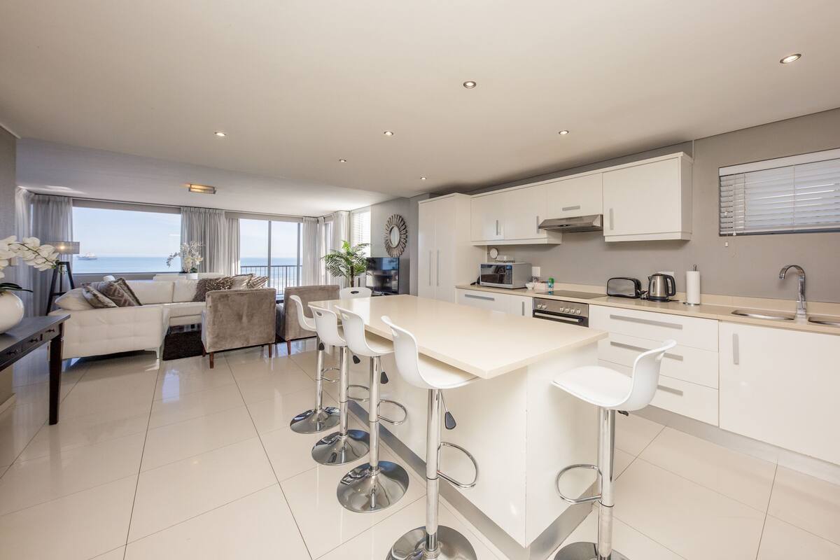 A modern kitchen with white cabinets, a central island with bar stools, and a view of the sea through large windows.