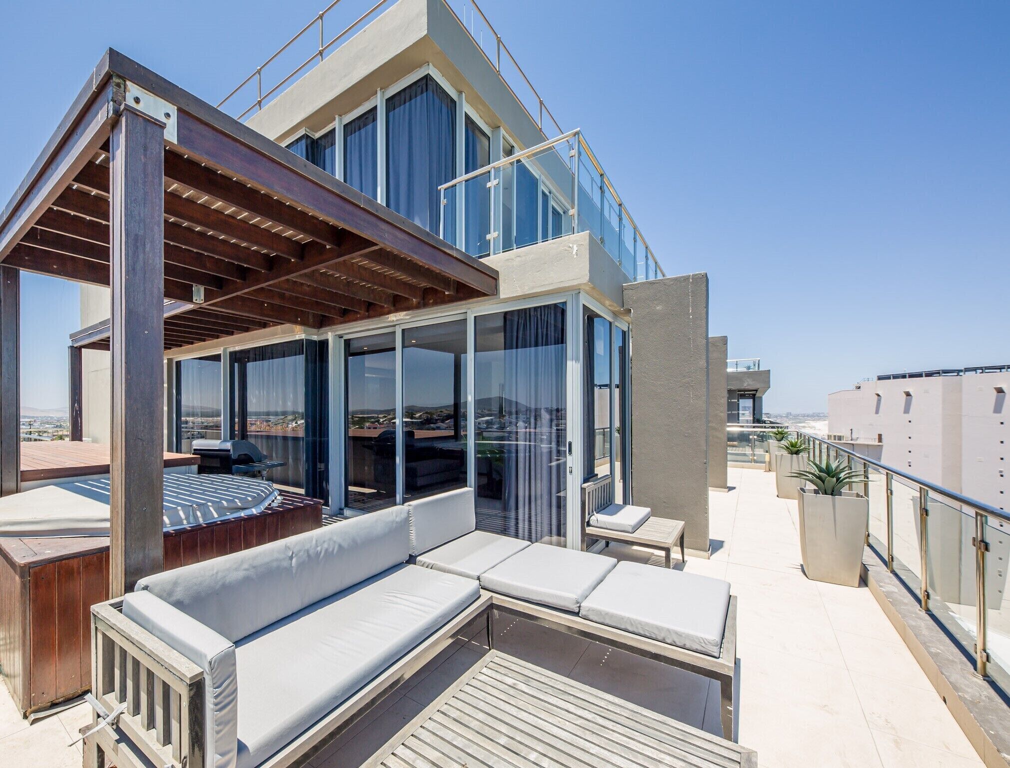 Una terraza en la azotea con una pérgola de madera, zona de estar y puertas de cristal que dan a un espacio cubierto.