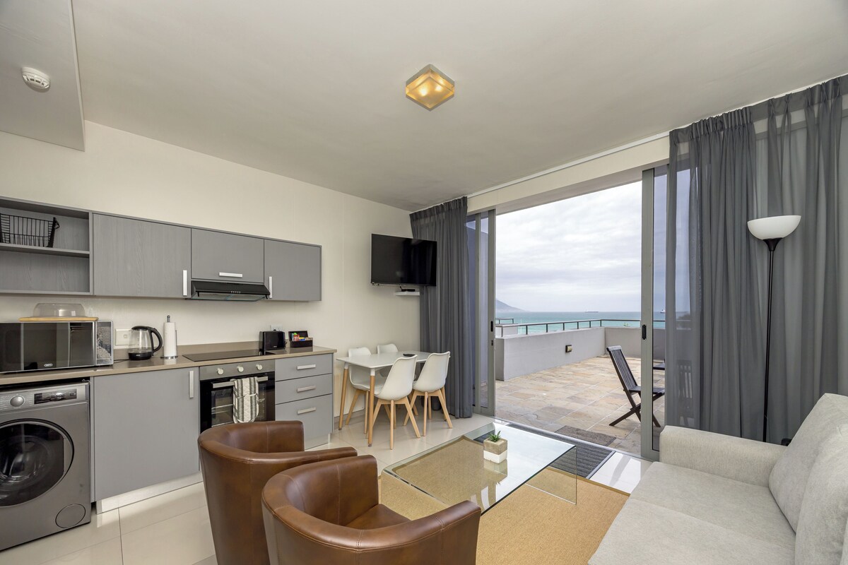 A modern kitchen with grey cabinets, a microwave, and a coffee maker. There is a dining area with a table and chairs, and a balcony with a view of the sea.