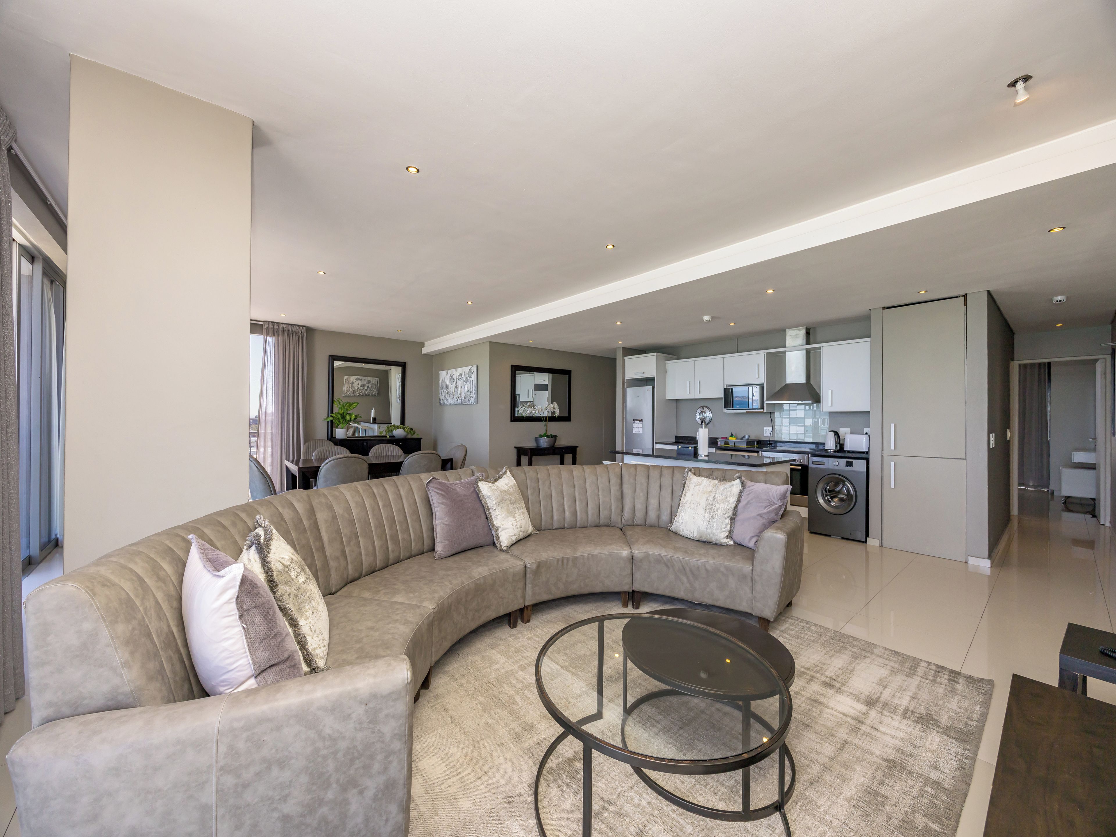 A modern living room with a curved sofa, a glass coffee table, and a kitchen in the background.