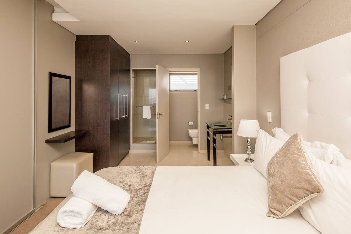 A modern hotel room with a large bed, a bedside table, a nightstand, a mirror, and a shower area.