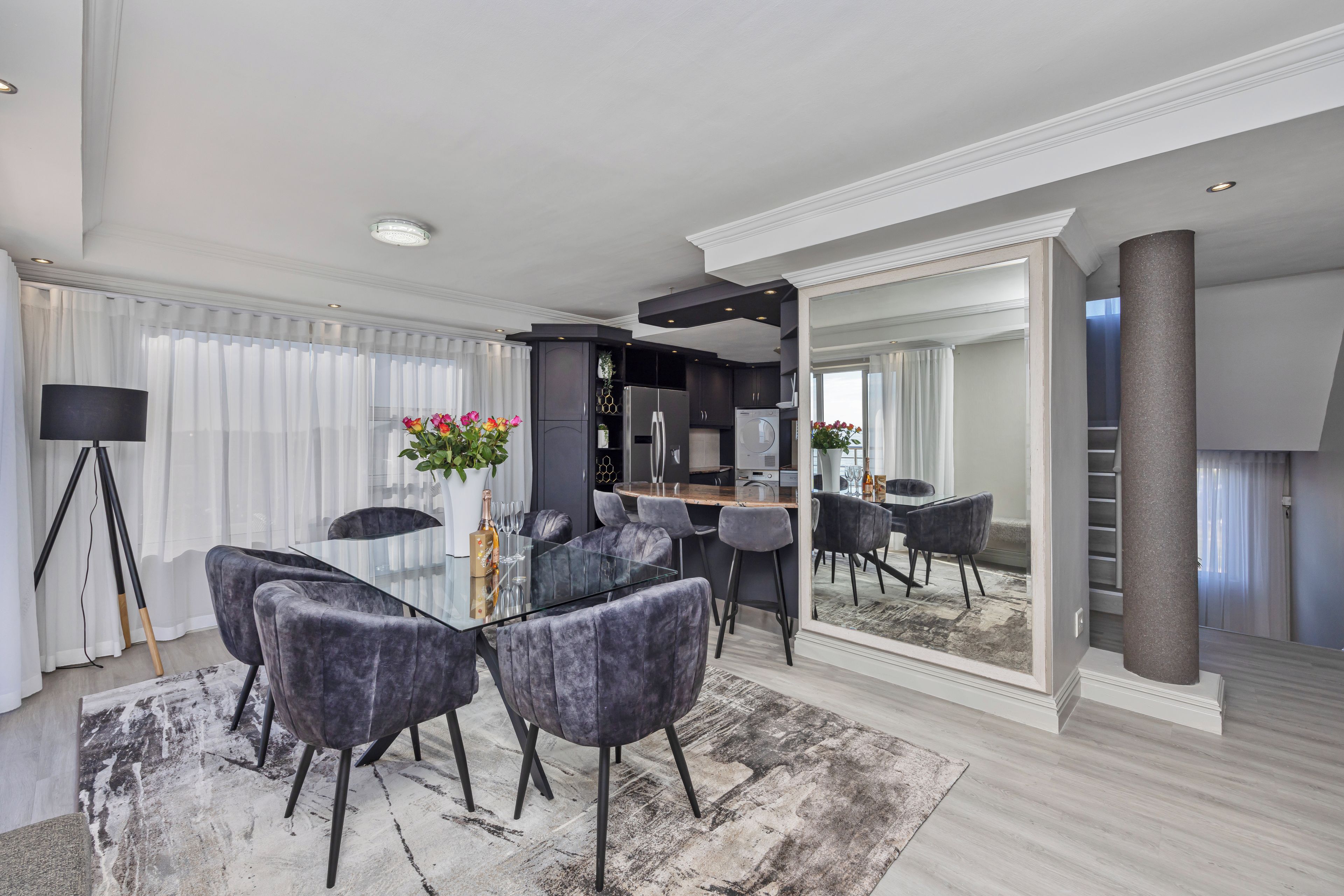 A modern living room with a glass dining table, grey upholstered chairs, a large mirror, and a dark kitchen area.