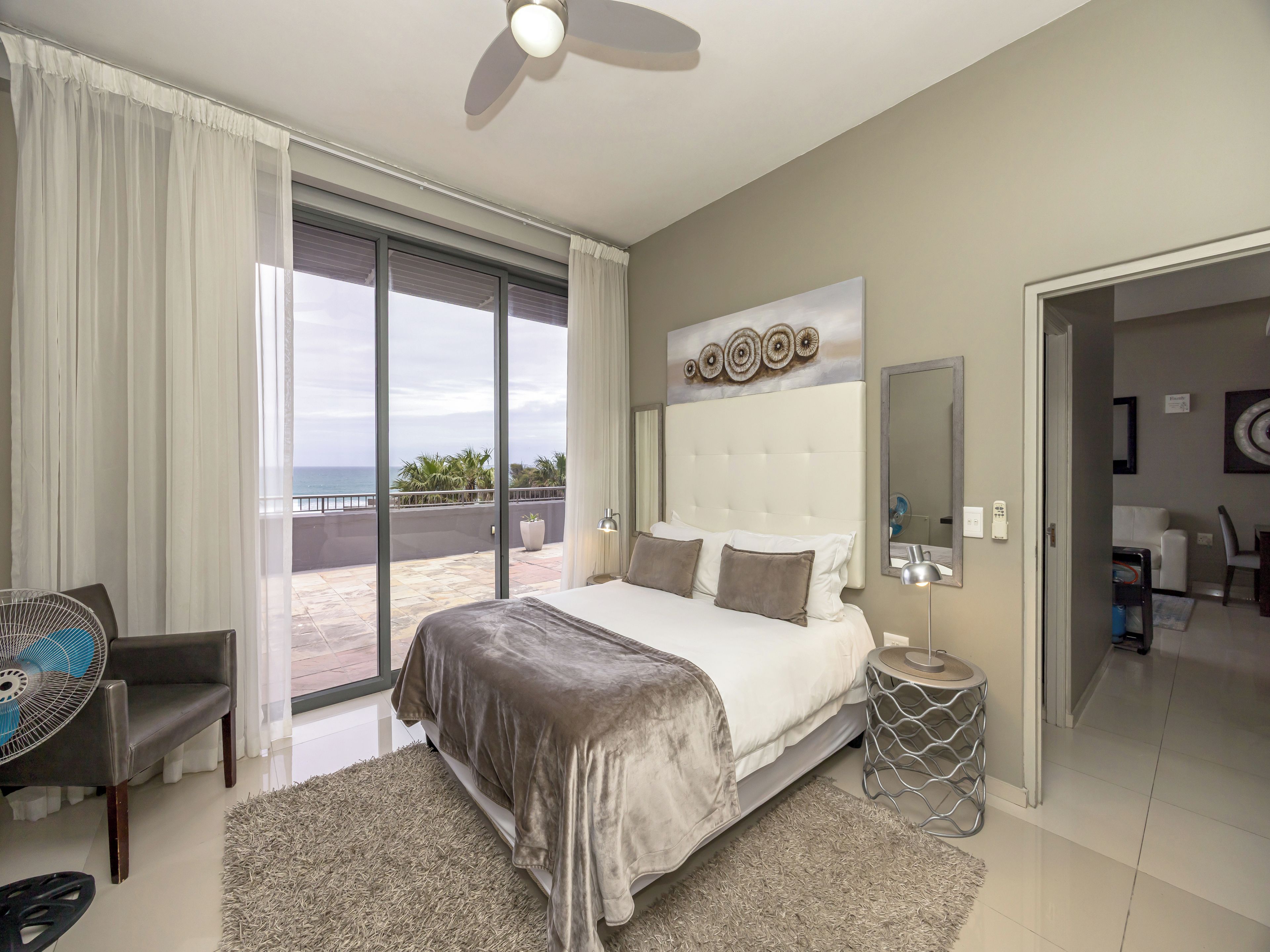 A hotel room with a large bed, a ceiling fan, a view of the ocean, and a balcony.