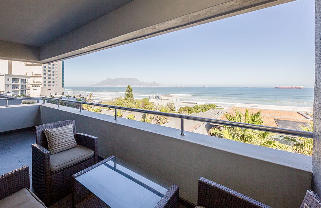 A balcony with wicker furniture and a glass table overlooking a beach and ocean.