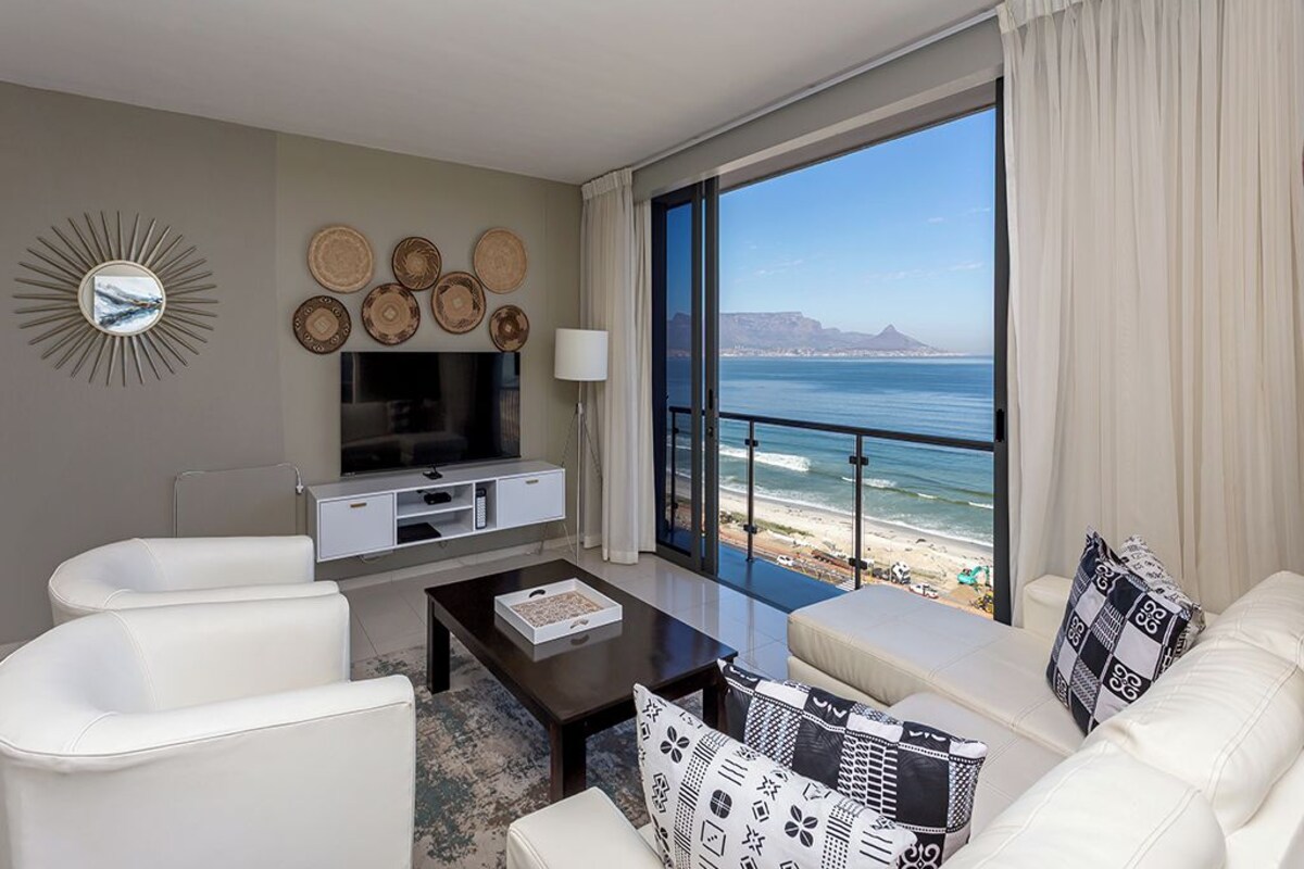 A modern living room with a large window overlooking a beach, a flat-screen TV, and a seating area with a coffee table.