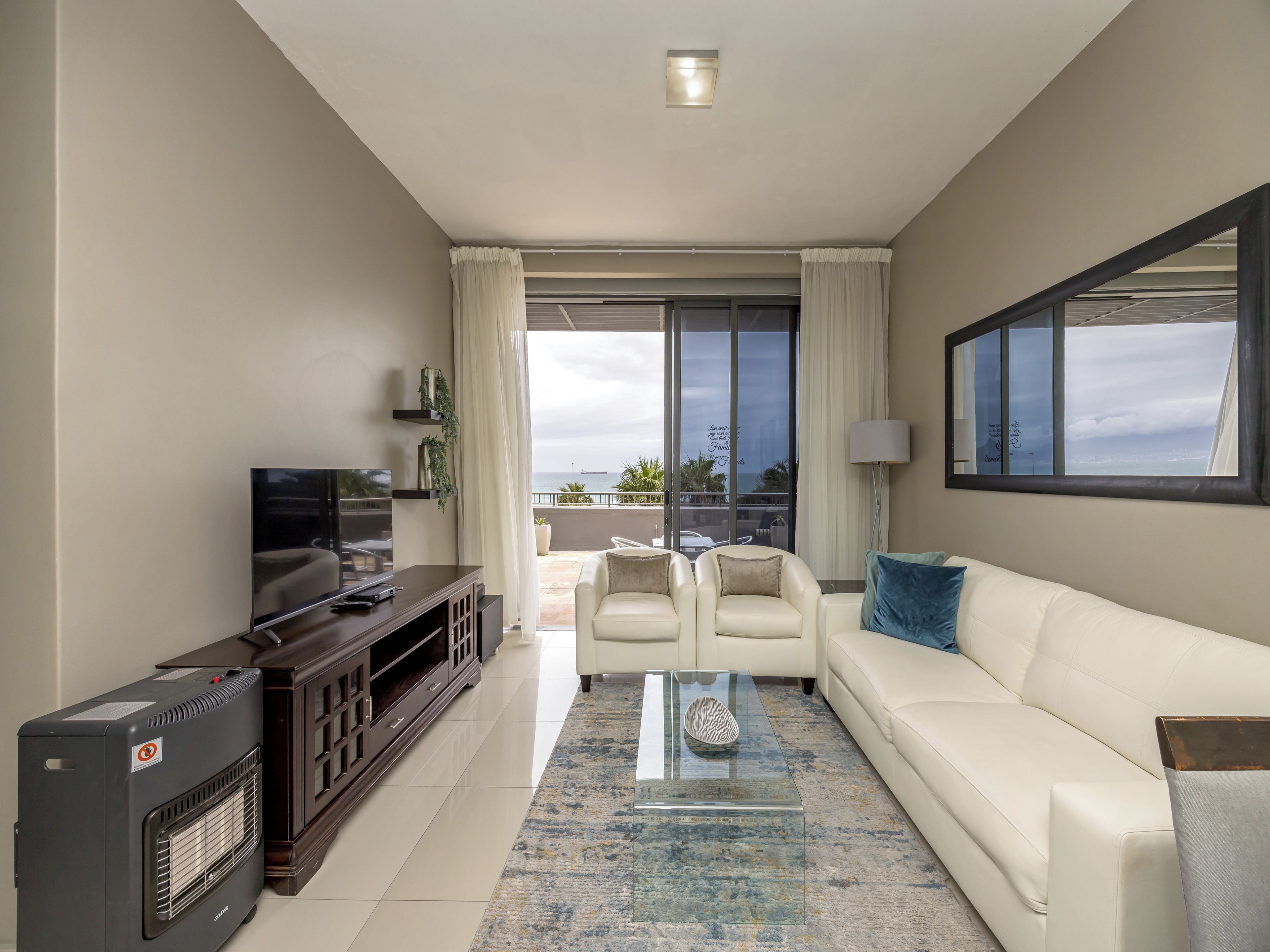 A modern living room with a flat-screen TV, a sofa, a coffee table, and a view of the sea through large windows.