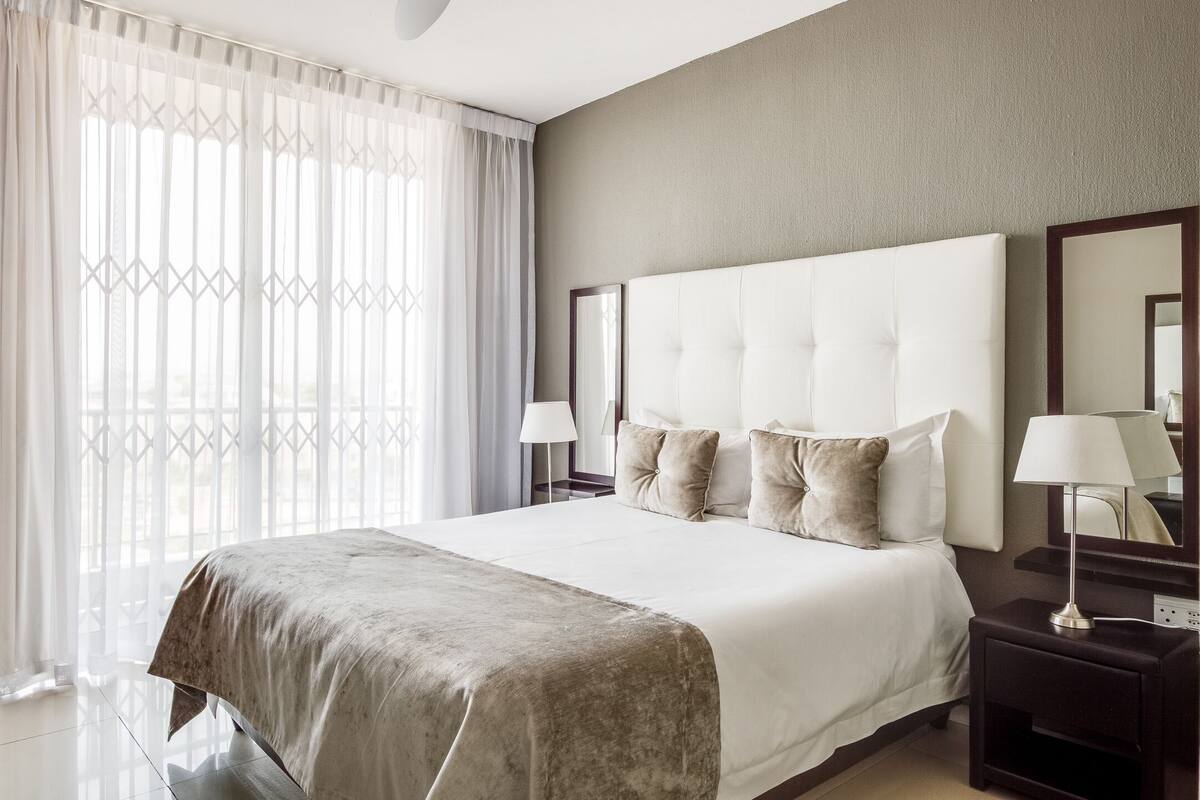 A hotel room with a large bed, two bedside tables, a mirror, and a window with sheer curtains.