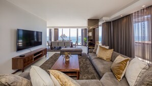A modern living room with a flat-screen TV, a wooden coffee table, and a large sofa. There is a dining area with a table and chairs, and a view of the ocean through large windows. - Infinity Self-Catering Apartments (Cape Town)