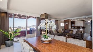 A modern living room with a wooden dining table, a sofa set, and a large window offering a view of the city.
