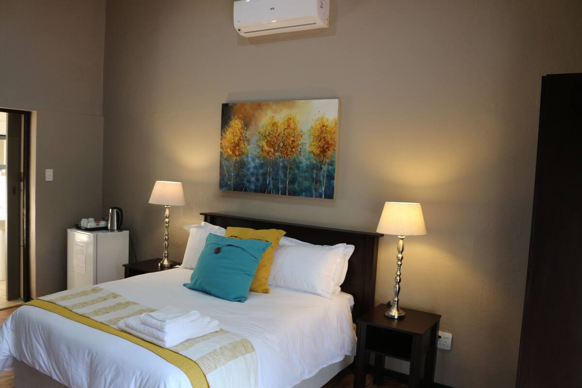 A neatly made bed with white and yellow bedding, two bedside lamps, a small table with a lamp, and a painting of trees on the wall.