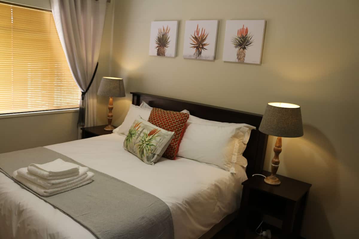 A neatly made bed with a gray throw blanket, white pillows, and a red patterned pillow. There are two wall-mounted lamps on either side of the bed, and three framed botanical prints above it.