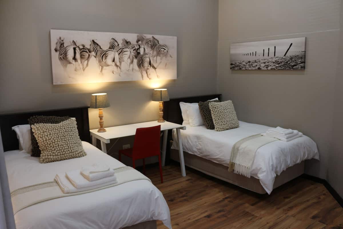 A hotel room with two single beds, a desk, two chairs, and two wall-mounted lamps.