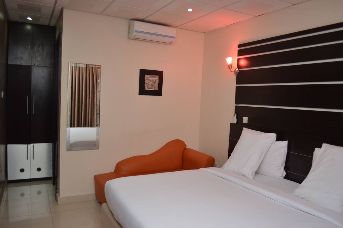 A hotel room with a bed, a wardrobe, a window with curtains, and a wall-mounted air conditioner.