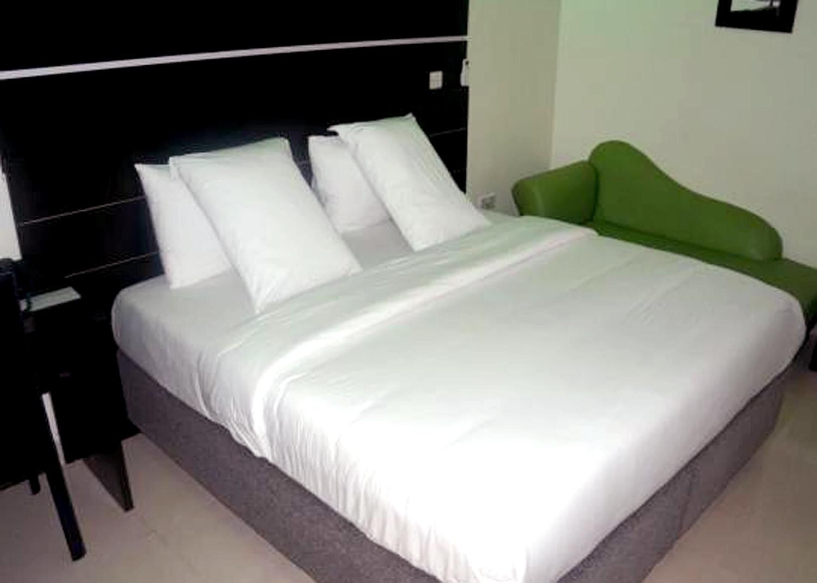 Una cama bien hecha con sábanas y almohadas blancas, un cabecero tapizado en verde y una mesita de noche de madera oscura.