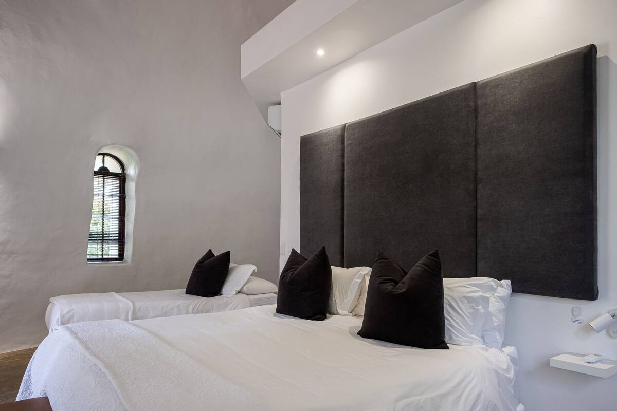 A modern hotel room with two beds, a large dark headboard, a small window, and a bedside table with a lamp.