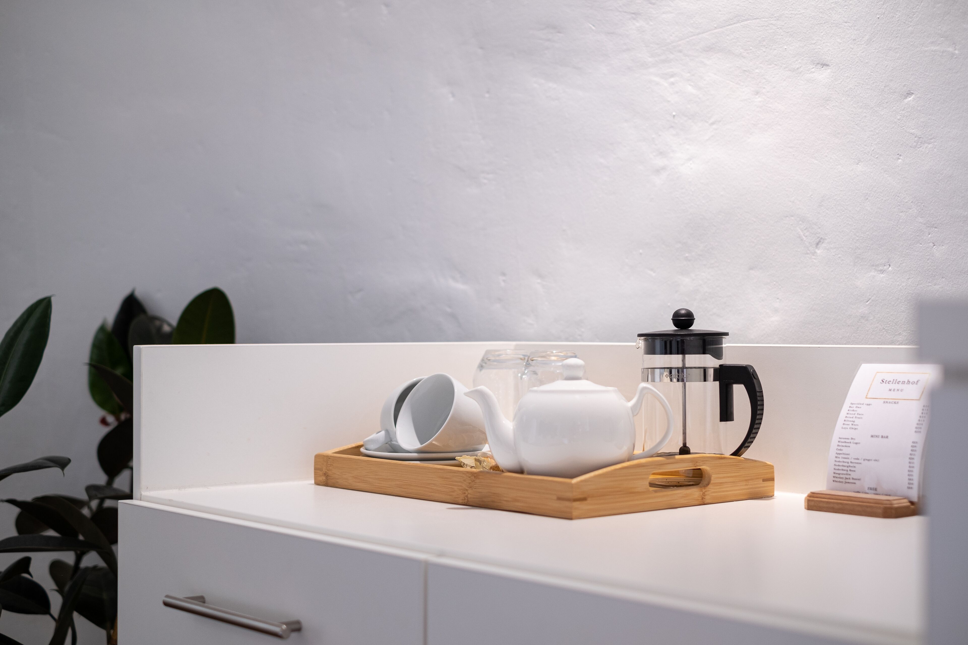 A white countertop with a wooden tray holding a white teapot, cups, and a coffee maker.
