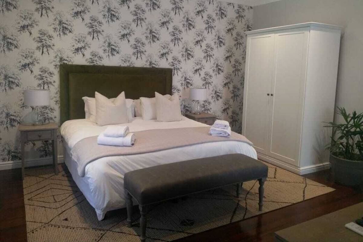 A neatly made bed with pillows and a folded towel, a bedside table with a lamp, a large wardrobe, and a potted plant.