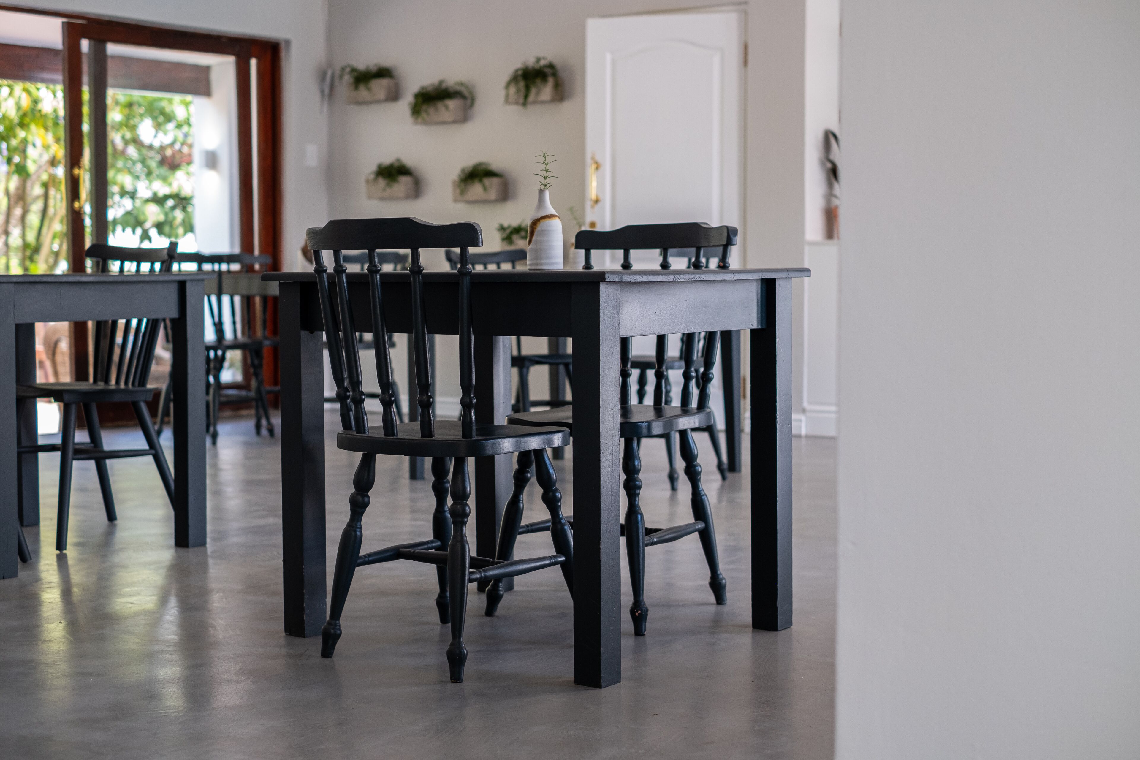 A dining area with a dark wooden table and chairs, a white vase with a plant, and a view of a door and wall-mounted planters.