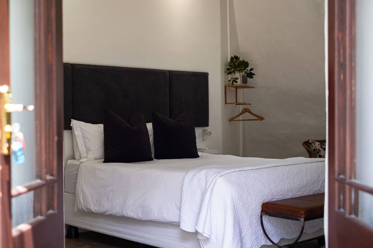 A neatly made bed with white bedding and two black pillows, a bedside table with a plant, and a wooden bench.