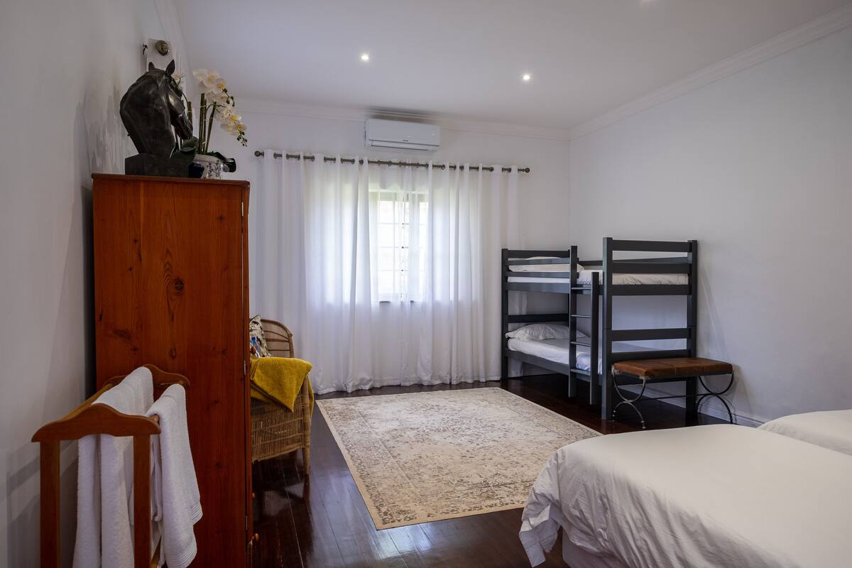 A bedroom with a bunk bed, a wooden wardrobe, a small table with a yellow cloth, and a window with white curtains.