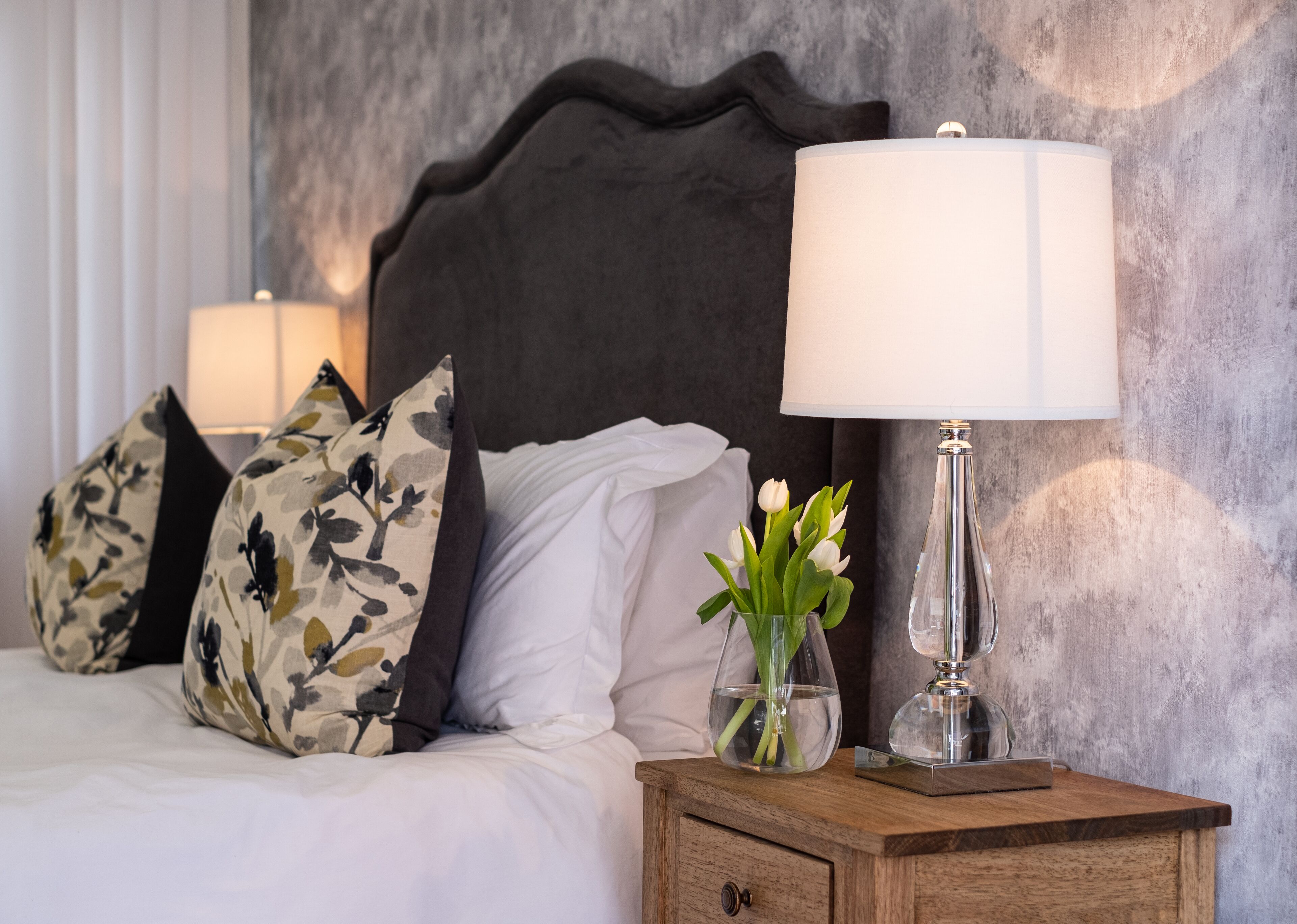 A neatly made bed with white linens, a dark headboard, and decorative pillows. A clear glass vase with white flowers is placed on a wooden nightstand beside the bed.