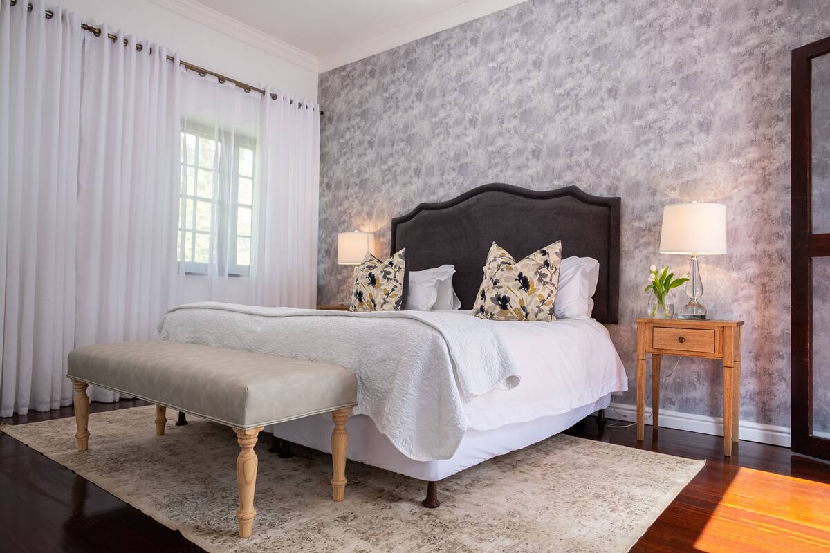 A bedroom with a large bed, a bench, two bedside tables with lamps, and a window with curtains.