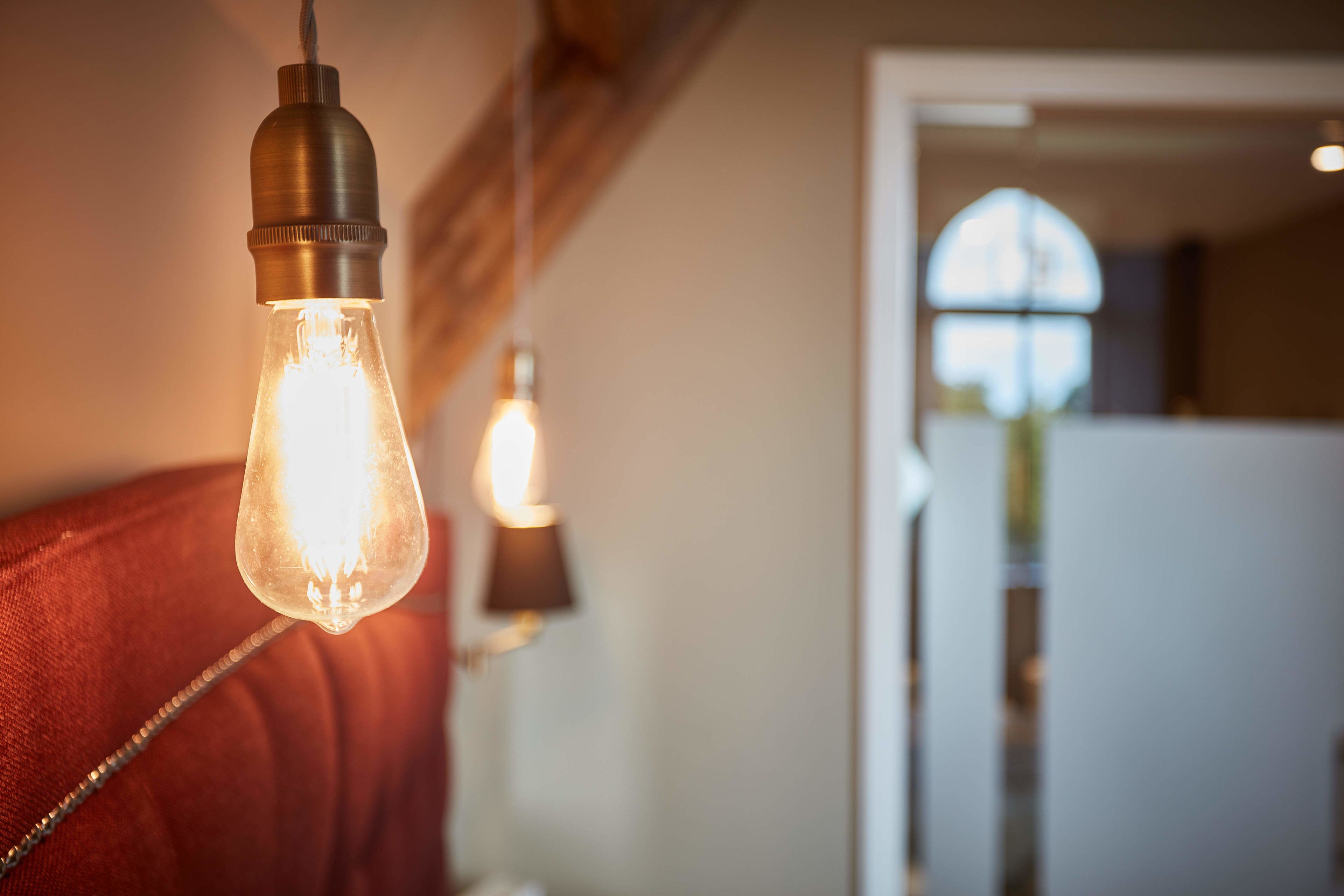 En närbild på en hängande glödlampa med en synlig glödtråd, en röd klädd sänggavel och en suddig bakgrund som antyder ett interiört utrymme.