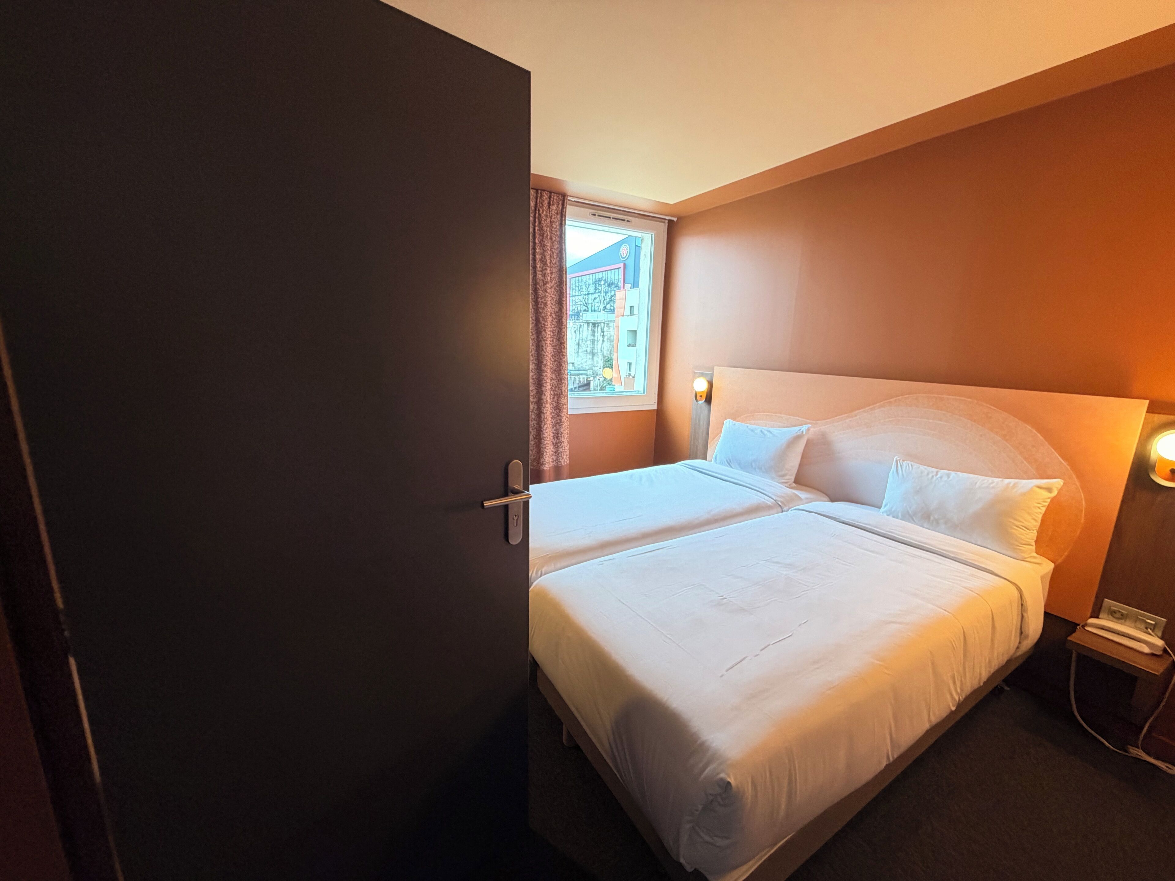 Une chambre d’hôtel avec un lit, une table de chevet, une lampe et une fenêtre avec des rideaux.