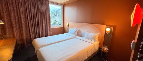 Habitación de hotel con cama doble, un escritorio, una silla, una ventana con cortinas y una lámpara de pared.