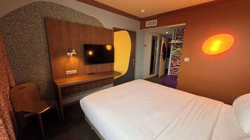 Een hotelkamer met een bed, een bureau met een televisie, een spiegel en een wandlamp.