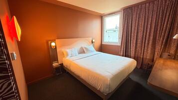 A hotel room with a bed, bedside table, lamp, and a view of the city through a window.