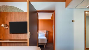 A hotel room with a bed, a TV mounted on the wall, a wooden cabinet, and a door leading to another room. - ibis Styles Alès Centre Cévennes (Ales)