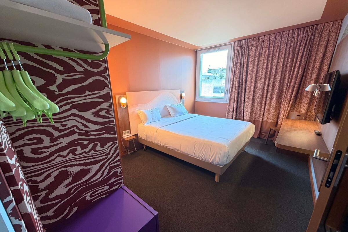 Une chambre d’hôtel comprenant un lit, un bureau, une chaise, une armoire, un portant à vêtements, une fenêtre avec des rideaux et un téléviseur fixé au mur.