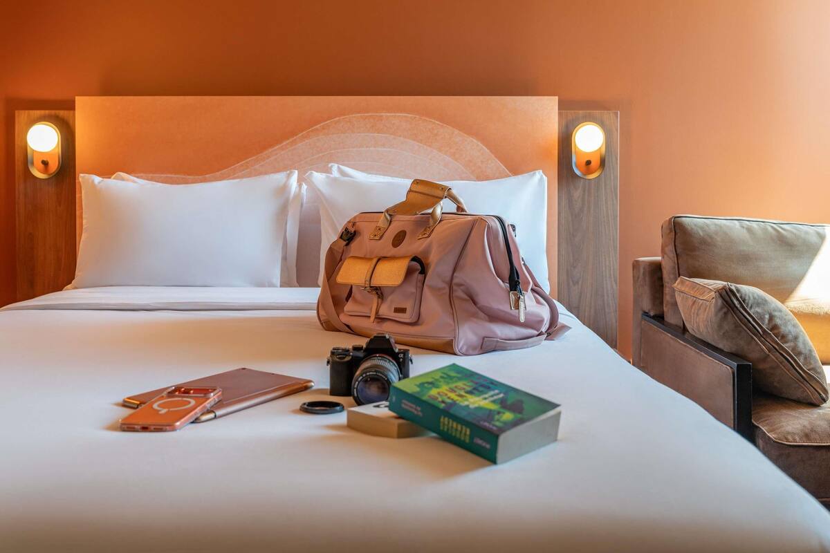 Un lit bien fait, avec des oreillers, un appareil photo, un livre et un sac posés dessus.