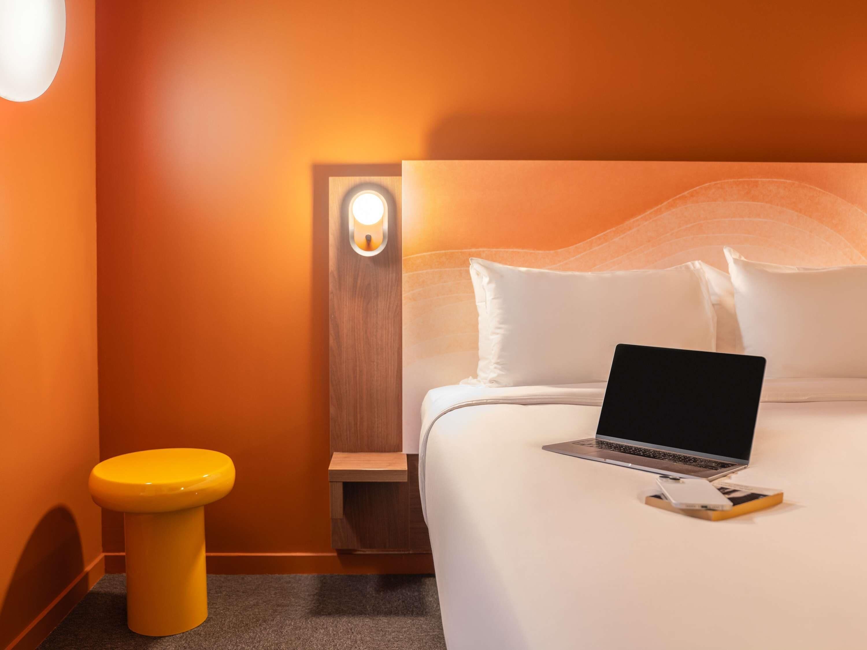A hotel room with a bed, a laptop, a yellow stool, and a nightstand.
