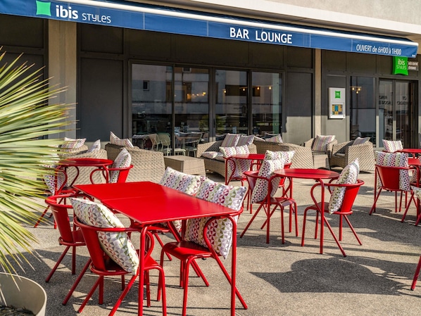 Un espace extérieur aménagé avec des tables et des chaises rouges, une enseigne indiquant la présence d’un bar, et une grande fenêtre.