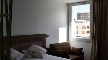 Een hotelkamer met een bed, een stoel, een raam met gordijnen en uitzicht op gebouwen.