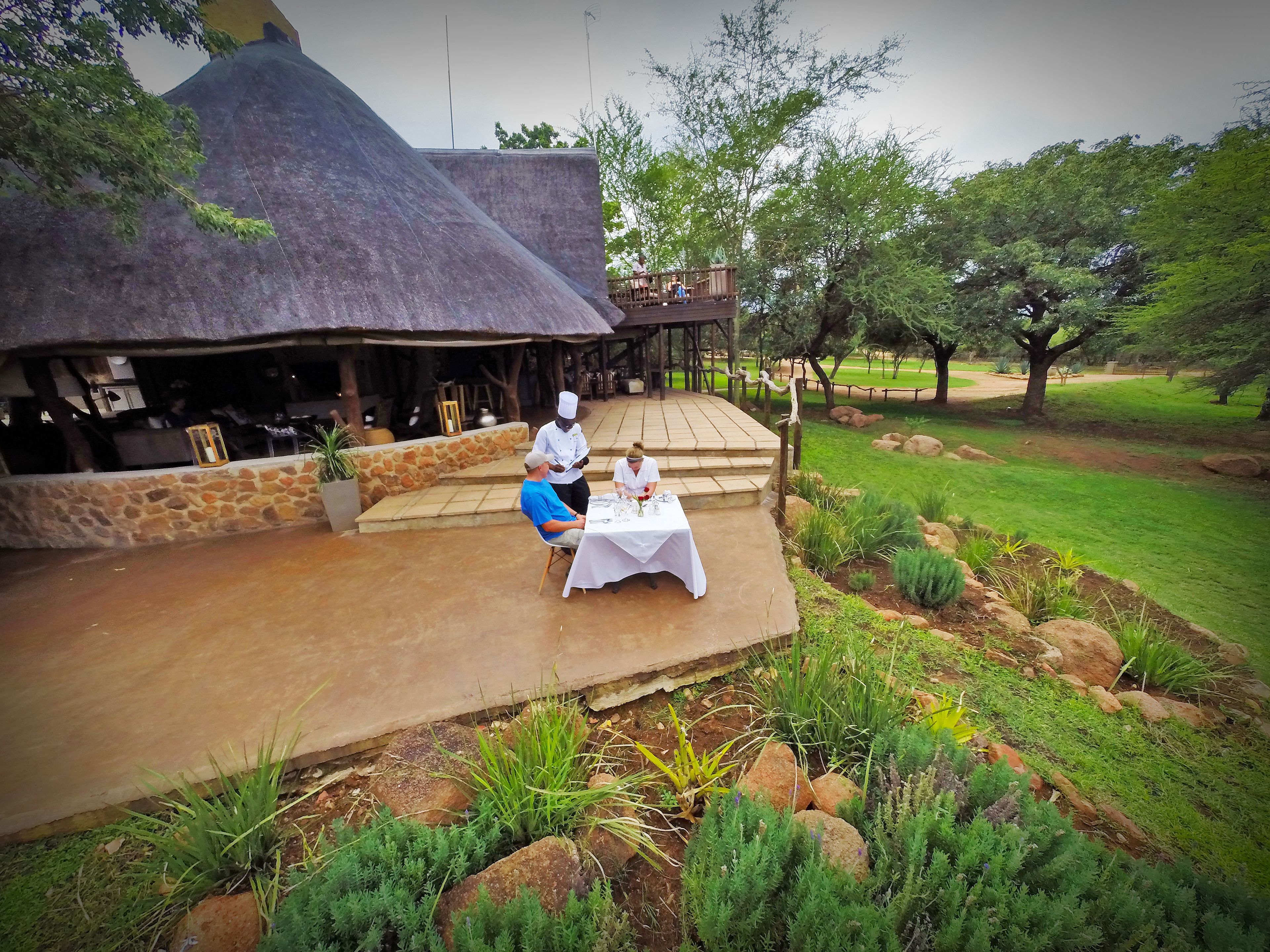 An outdoor dining area with a thatched-roof structure, stone walls, and a garden with rocks and greenery.