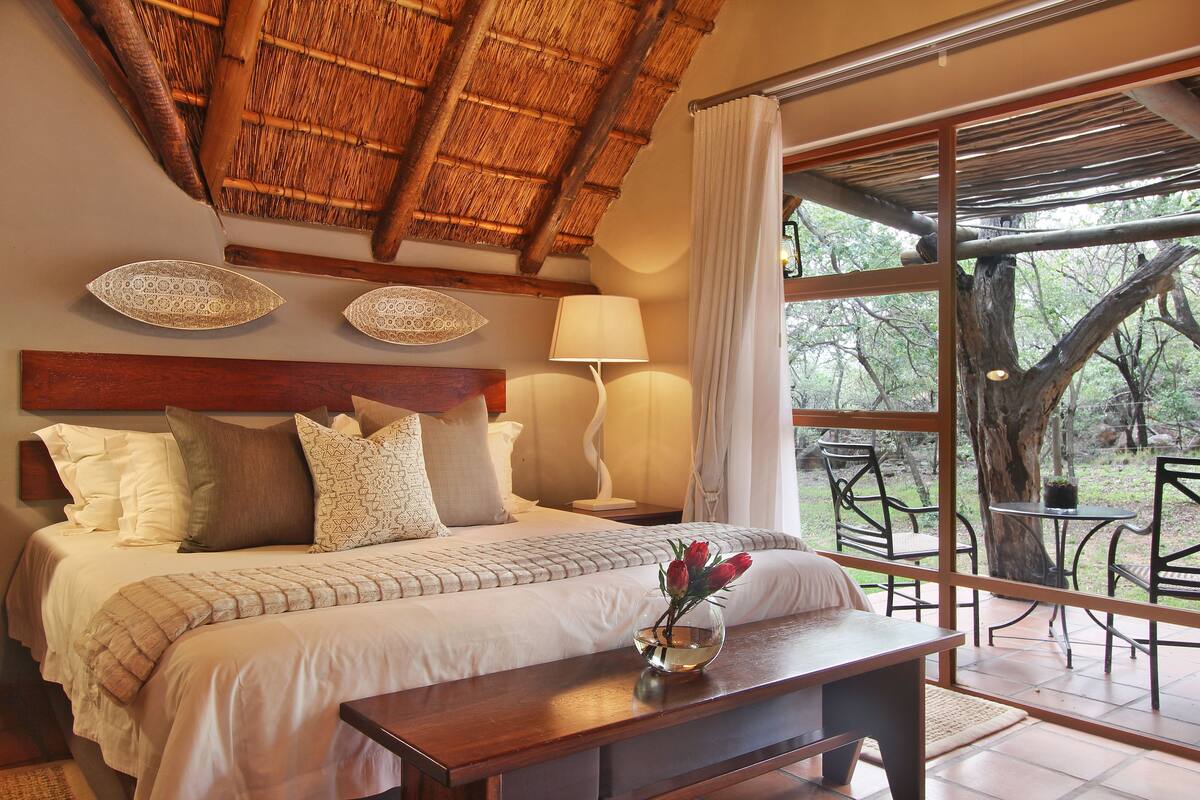 A bedroom with a large bed, a bedside table with a vase of flowers, a nightstand with a lamp, and a view of the outdoors through a glass door.