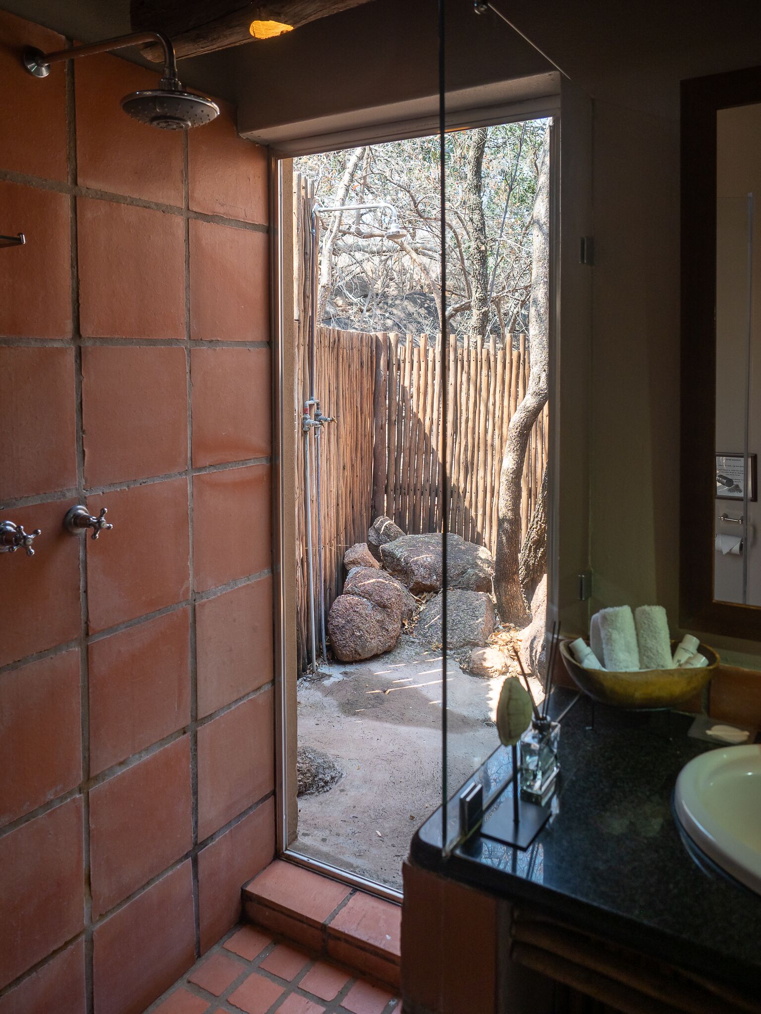 Een badkamer met een douche, een wastafel en uitzicht op een tuin met rotsen en een houten hek.