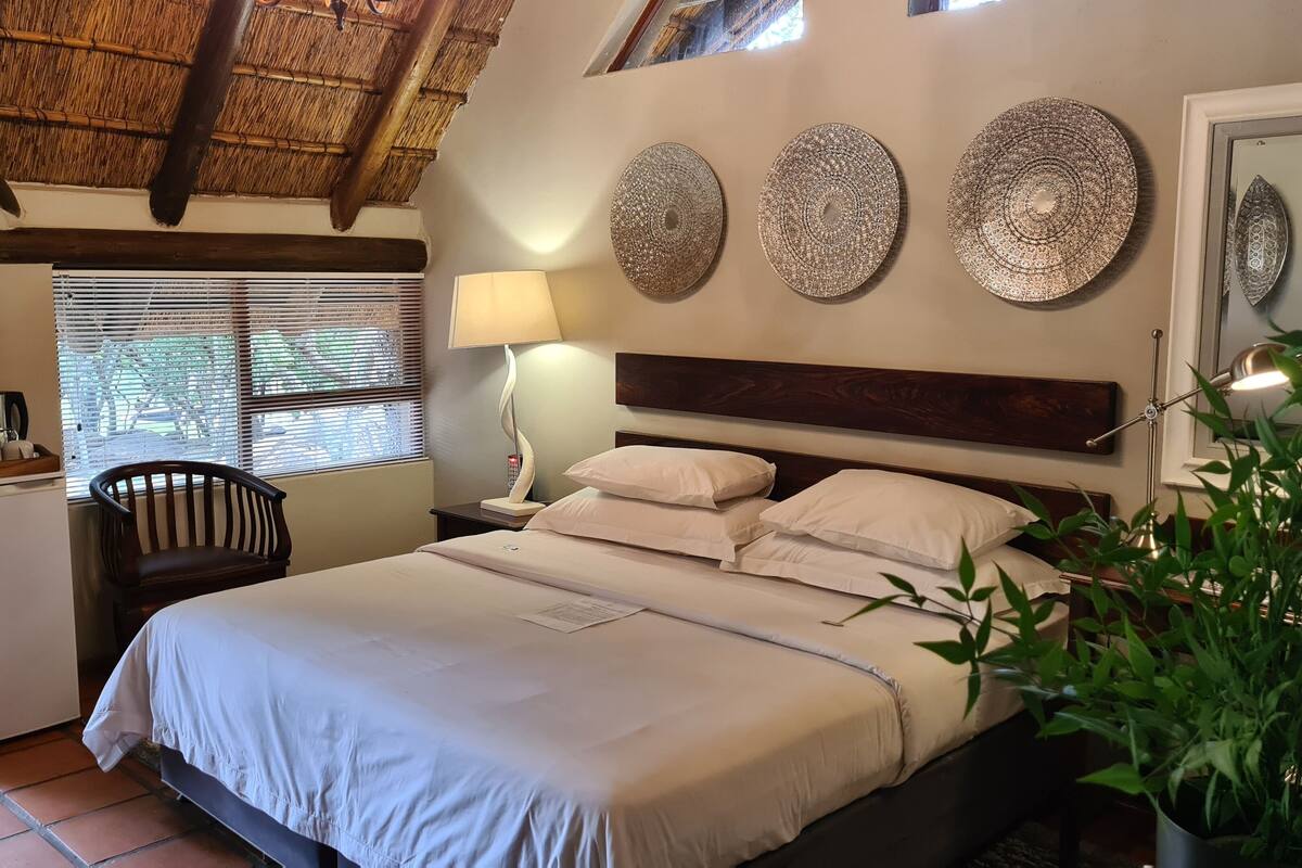 A bedroom with a bed, a desk, a chair, a refrigerator, and a potted plant.