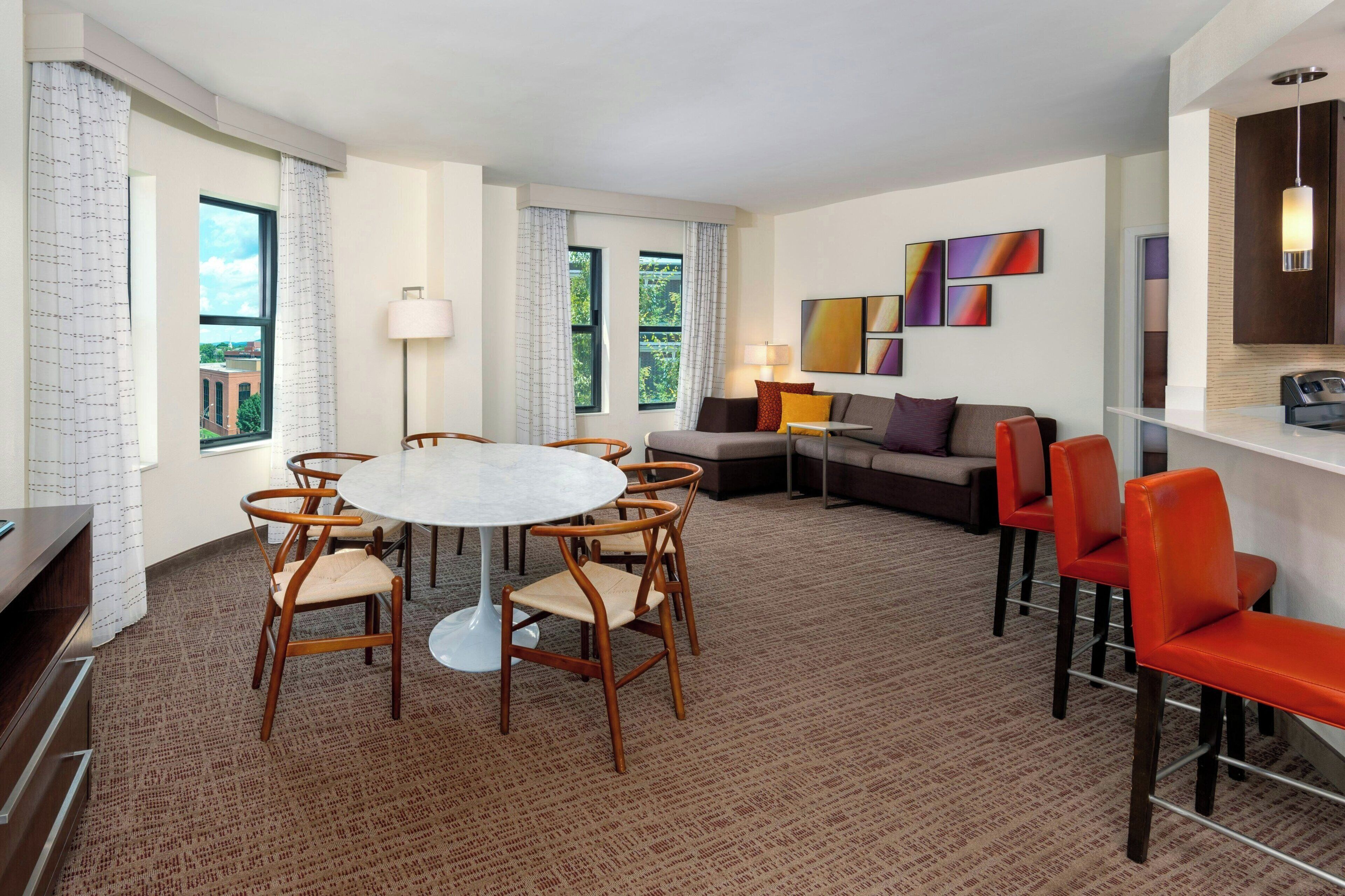 A modern hotel room with a round dining table, wooden chairs, a sofa, and a TV.