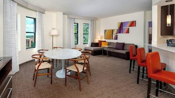 A modern hotel room with a round dining table, wooden chairs, a sofa, and a TV.