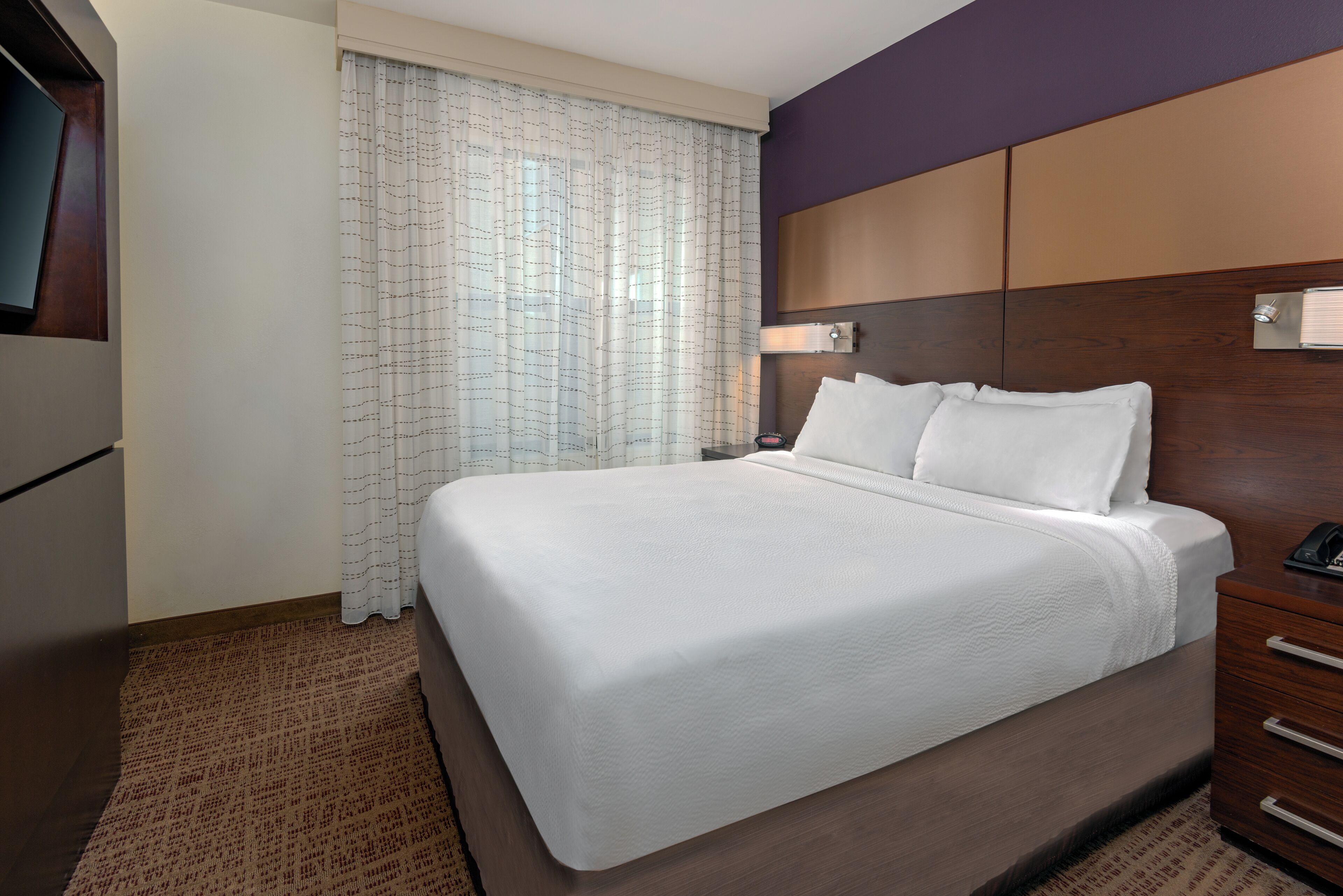 A hotel room with a large bed, wooden headboard, a bedside table with a lamp, and a window with curtains.
