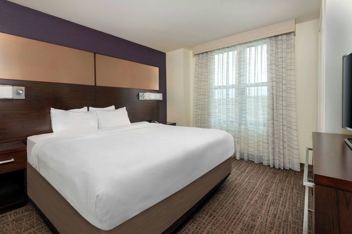 A hotel room with a large bed, a wooden headboard, a flat-screen TV, and a window with curtains.