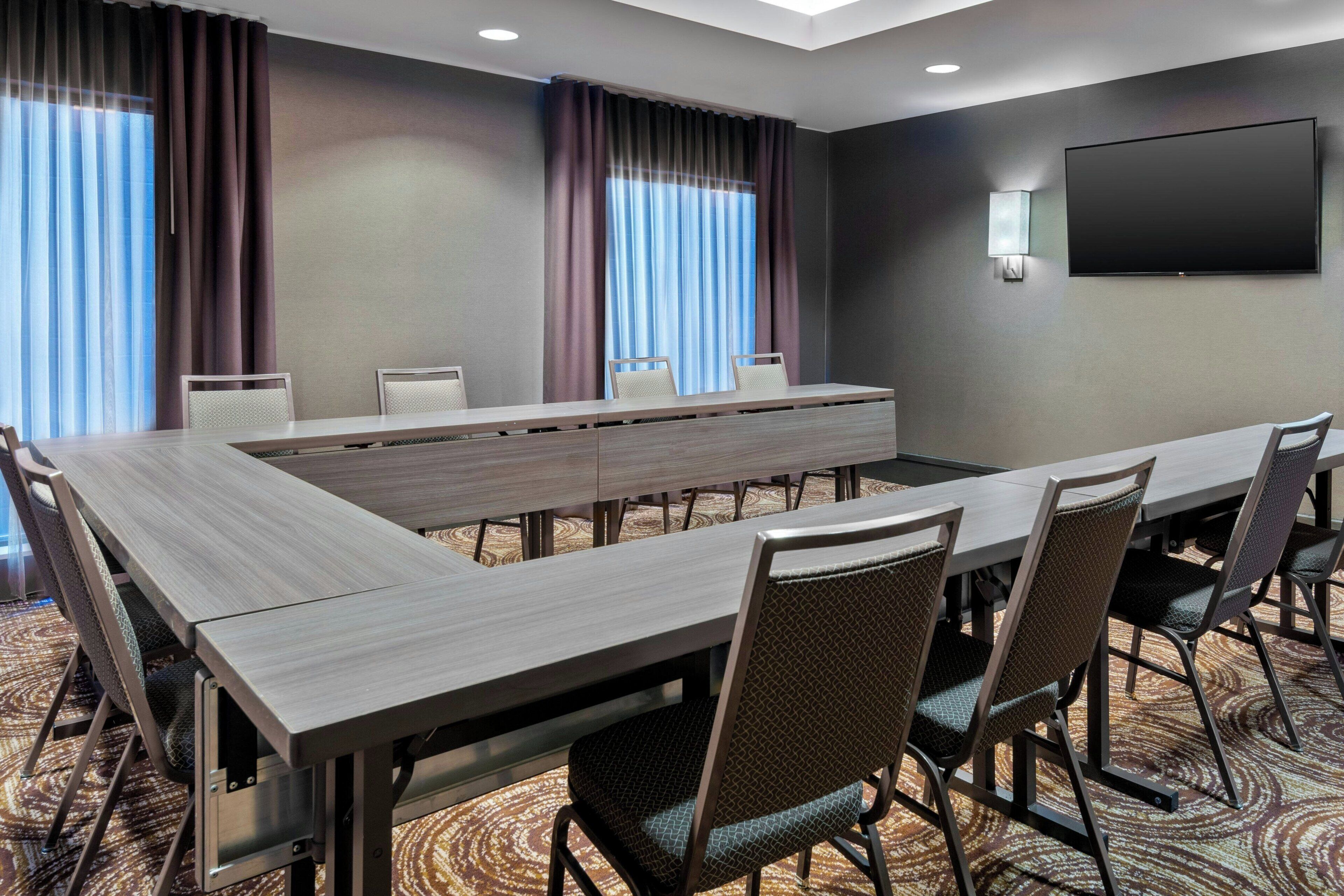 A conference room with a long table, chairs, a flat-screen TV, and curtains.